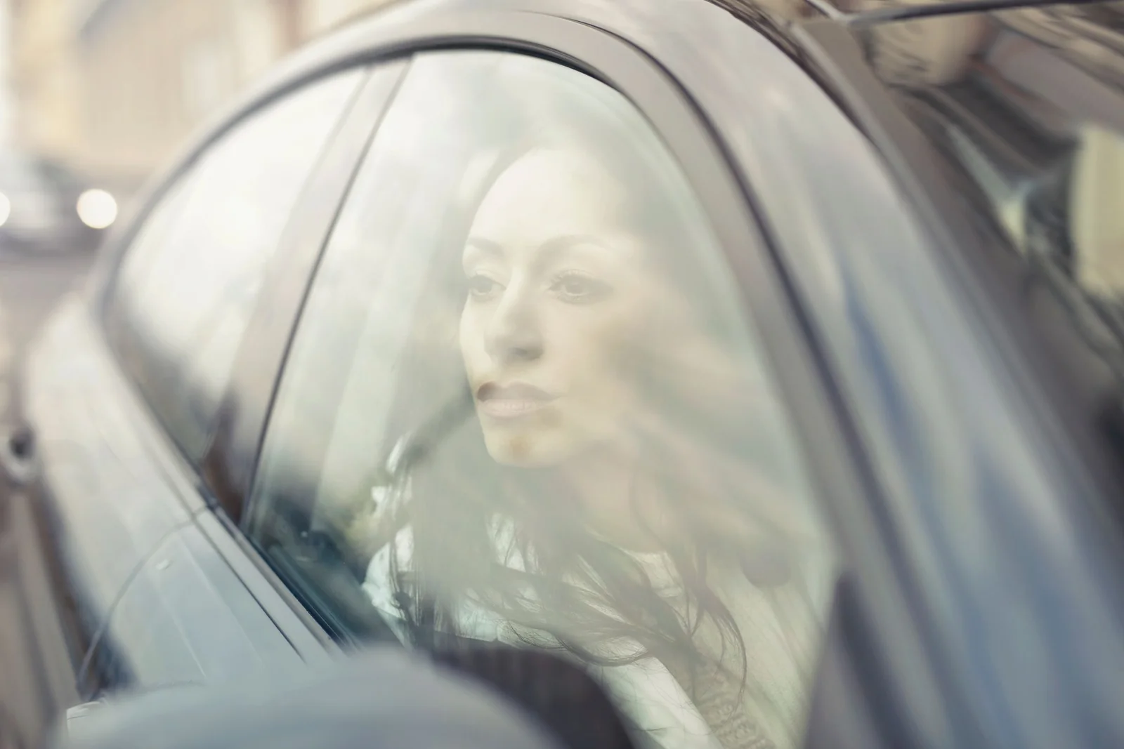 A woman reflects while sitting inside a car, looking through the window with a thoughtful expression.